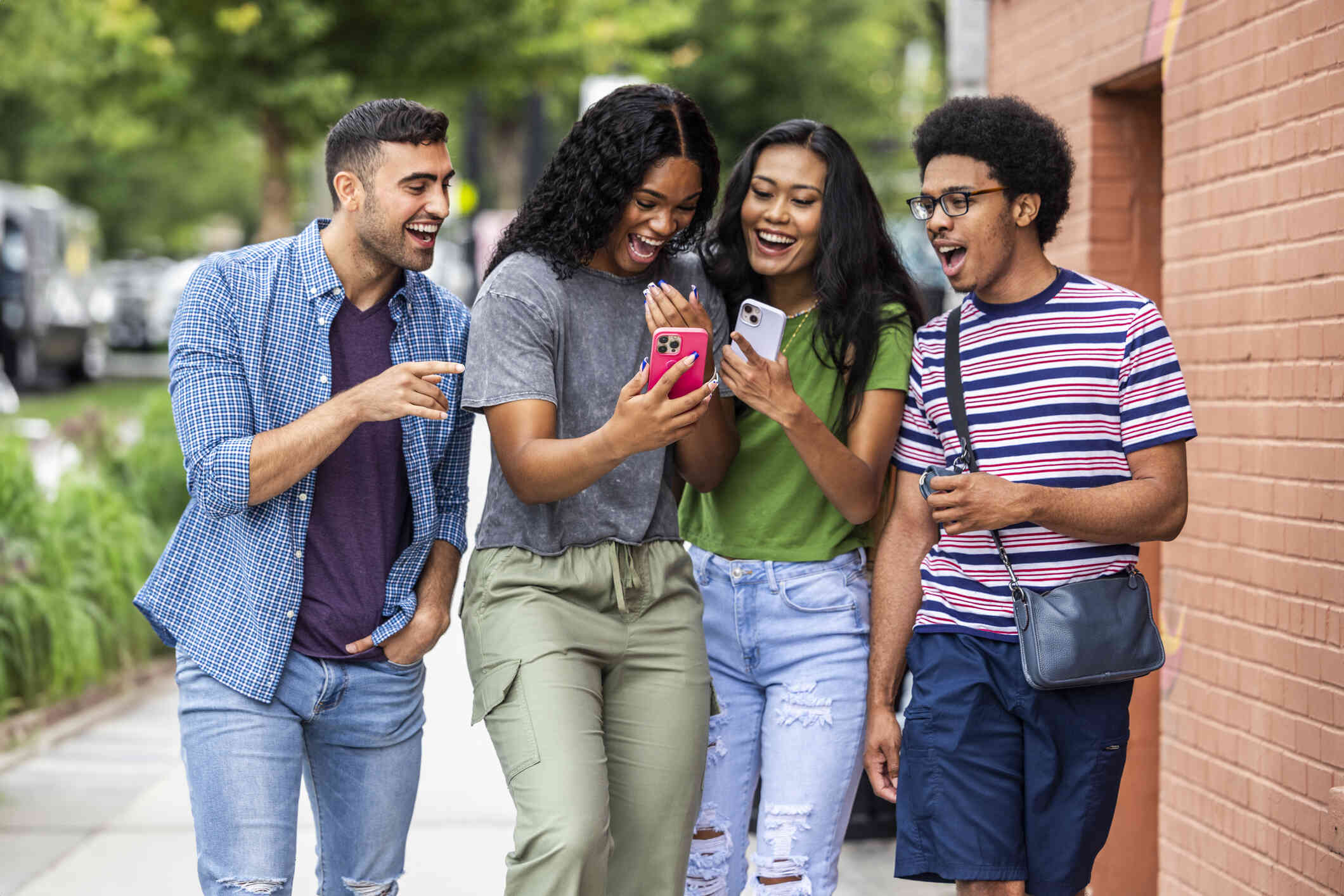 A group of adults stand together outside and look at a cellphone while smiling and laughing.